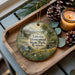 A wooden tray with a decorative ornament featuring a scenic landscape of a cabin and a waterfall, surrounded by pine cones and a lit candle.