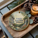 A decorative ornament featuring a scenic landscape of Loggy State Park, Arkansas, with a waterfall and pine trees. The ornament is placed on a wooden tray, accompanied by pine cones and a lit candle.