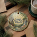 A round ornament with a scenic landscape of Logoly State Park, Arkansas, featuring a waterfall and pine trees, is displayed on a wooden surface alongside a cup of coffee and some papers.