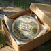 A decorative ornament in a box, featuring a scenic landscape with mountains, water, and greenery.