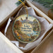 A decorative ornament in a box, featuring a scenic landscape with mountains, a river, and wildlife, including a deer.