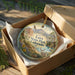 A decorative ornament in a box, featuring a scenic landscape with mountains, trees, and wildlife, including a deer.