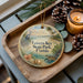 A decorative ornament with a tropical beach scene, featuring a bird, palm trees, and a body of water, is displayed on a wooden tray alongside pine cones and a lit candle.