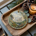 A wooden tray with a decorative ornament featuring a scenic landscape of the state park in Connecticut, accompanied by pine cones and a lit candle.