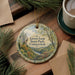 A decorative ornament with a scenic landscape of a river and forest, featuring the text "Lovers Leap State Park Connecticut" on it, placed on a wooden surface alongside a cup of coffee and some pine branches.
