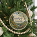 A round ornament hanging on a Christmas tree, featuring a scenic landscape with a river and mountains.