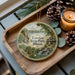A decorative ornament featuring a scenic landscape of Lowden State Park, Illinois, with a candle and pine cones on a wooden tray.