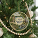 A round ornament hanging on a Christmas tree, featuring a scenic landscape with a lake, trees, and a cabin.