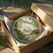 A decorative ornament in a box, featuring a scenic landscape with a waterfall and a cabin, with the text "Lowden State Park, Illinois" printed on it.