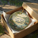 An oval-shaped ornament with a scenic landscape of a pond, trees, and a boat, displayed in a cardboard box with a gold string.