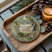 A decorative ornament with a scenic image of a park, featuring a bridge, trees, and flowers, is displayed on a wooden tray alongside pine cones and a lit candle.