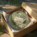 A decorative ornament in a brown box, featuring a scenic landscape with a bridge and greenery, with the text "Macedonia Brook State Park, Connecticut" printed on it.