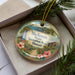 A decorative ornament in a box, featuring a tropical landscape with palm trees, flowers, and a waterfall.