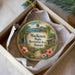 A circular ornament with a tropical landscape design, featuring a waterfall, palm trees, and flowers, is displayed in a box.