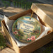 A decorative ornament in a brown box, featuring a tropical landscape with a waterfall and flowers, is placed on a wooden surface.