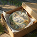 A circular ornament with a scenic landscape and text, placed in a cardboard box with tissue paper.