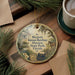 A circular ornament with a vintage-style illustration of a historic park, featuring a horse, typewriter, and other elements, is displayed on a wooden surface alongside a cup of coffee and some papers.