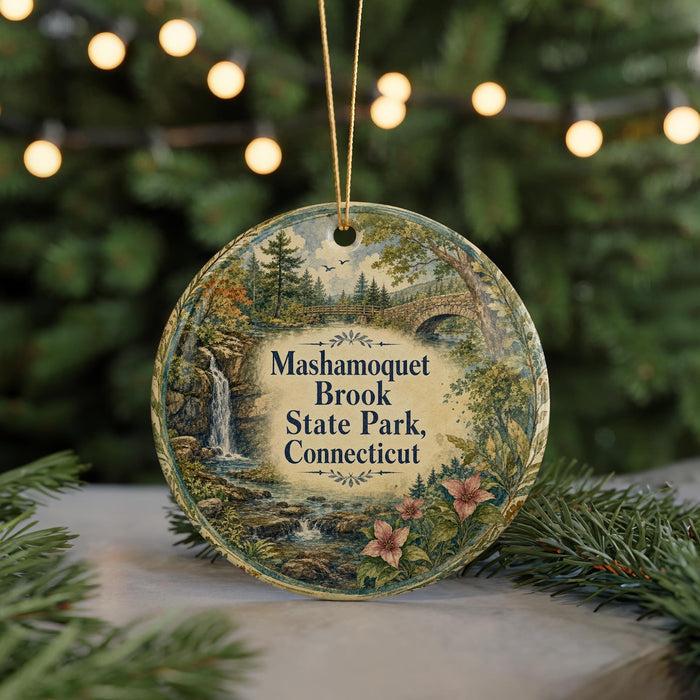 A round ornament with a scenic landscape of a waterfall, bridge, and trees, surrounded by greenery and placed on a surface with a blurred background of lights.