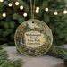 A round ornament with a scenic landscape of a waterfall, bridge, and trees, surrounded by greenery and placed on a surface with a blurred background of lights.