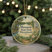 A circular ornament with a scenic landscape of a lake, trees, and a castle, hanging from a string.