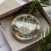 A decorative ornament with a scenic landscape featuring mountains, waterfalls, and a cabin, hanging from a string.