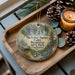 A decorative ornament featuring a scenic image of the Arthur Burney Falls Memorial State Park in California, with pine cones and a lit candle on a wooden tray.