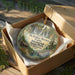 A decorative ornament in a wooden box, featuring a scenic landscape with a waterfall and a cabin, with the text "McArthur-Burney Memorial State Park, California" printed on it.