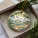 A decorative ornament in a box, featuring a scenic landscape with pine trees, a lake, and mountains.