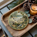 A decorative ornament featuring a scenic landscape of the state park in Iowa, with a candle and pine cones on a wooden tray.