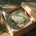 A decorative ornament in a wooden box, featuring a scenic landscape with a waterfall and flowers, with the text "McIntosh Woods State Park, Iowa" printed on it.