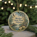 A circular ornament with a scenic image of a park, featuring a bird, a bridge, and flowers, is displayed in front of a blurred background of Christmas lights.