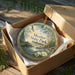 A round ornament in a box, featuring a scenic image of a park with a bridge and trees, with the text "Meather State Park, Alabama" printed on it.