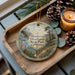 A decorative ornament featuring a scenic landscape of Mendocino Headlands State Park, California, with a lighthouse and pine trees, is displayed on a wooden tray alongside pine cones and a lit candle.