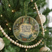 A round ornament hanging on a Christmas tree, featuring a forest scene with a deer and a cabin.