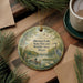A round ornament with a painting of ducks and a bird in a pond, surrounded by greenery, hangs on a wooden surface.