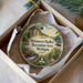 A round ornament with a scenic landscape and a boat on a lake, displayed in a box with a white tissue paper lining.