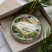 A decorative ornament with a scenic landscape of Metamora-Hadley Recreation Area, Michigan, featuring a cabin, a river, and a boat.