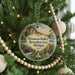 A circular ornament hanging on a Christmas tree, featuring a scenic landscape with a river, a cabin, and a waterfall.