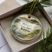 A decorative ornament in a box, featuring a scenic landscape with a lake, mountains, and a bird, along with the text "Milford State Park, Kansas".
