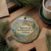 A decorative ornament with a scenic landscape of Millerton Lake, the State Recreation Area in California, featuring a lake, mountains, and pine trees. The ornament is hanging from a string and is placed on a wooden surface, with a cup of coffee and some papers nearby.