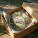A round ornament in a box, featuring an illustration of a park with a castle, trees, and a waterfall.