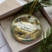 A decorative ornament in a box, featuring a scenic landscape with a bird, trees, and a castle-like structure.