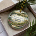A Minnesota Valley Recreation Area ornament is displayed in a box, featuring a scenic landscape with a bridge, pine trees, and wildlife.