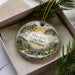 A decorative ornament in a box, featuring a scenic landscape with a bridge, trees, and flowers.