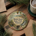 A decorative ornament featuring a scenic landscape with a waterfall, bridge, and pine trees, hanging on a wooden surface.