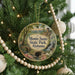 A round ornament hanging on a Christmas tree, featuring a scenic landscape with a bridge and a waterfall.