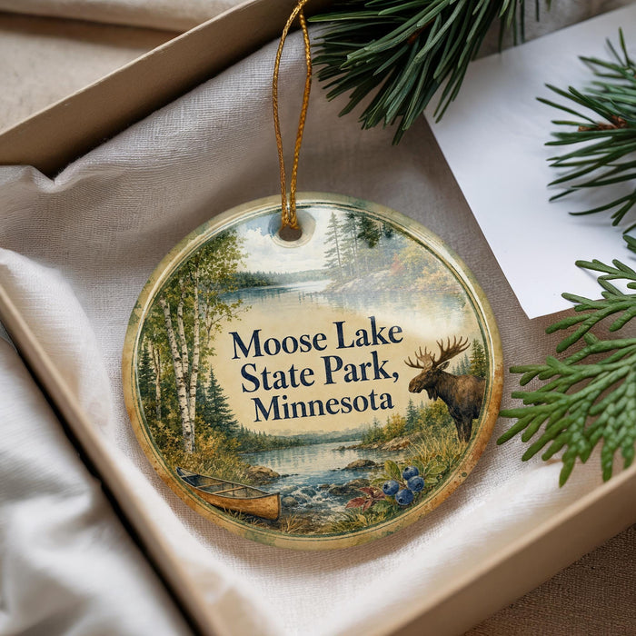 A circular ornament with a moose and pine trees, surrounded by greenery, is displayed in a box.