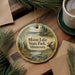 A circular ornament with a moose and a lake, surrounded by pine branches, sits on a wooden surface.