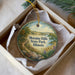 A decorative ornament in a white box, featuring a scenic landscape with a path, trees, and a body of water.