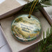 A decorative ornament in a box, featuring a scenic landscape with a path, trees, and a body of water.